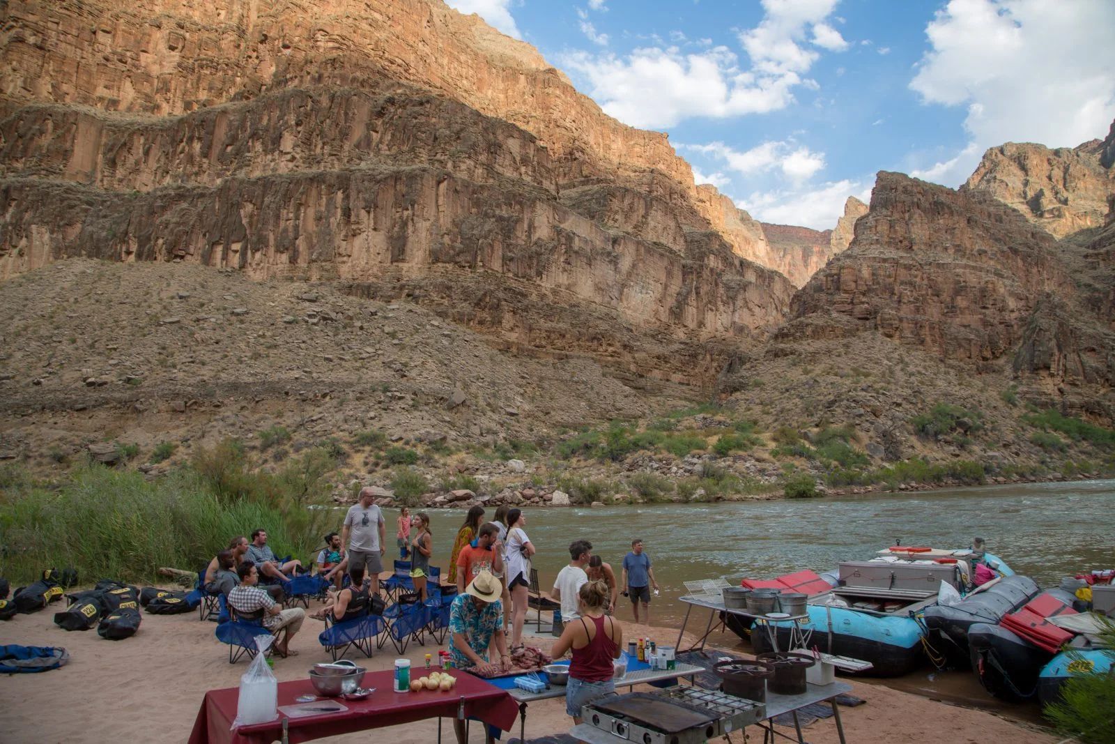 People gathered near a river with rafts and canyon walls in the background.