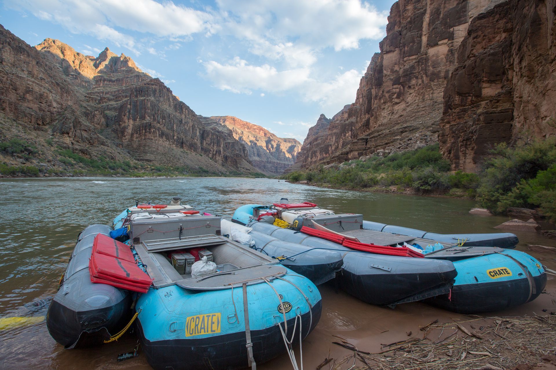 Rafts sit along a riverbank in a canyon. Brown cliffs rise on either side under a blue sky.
