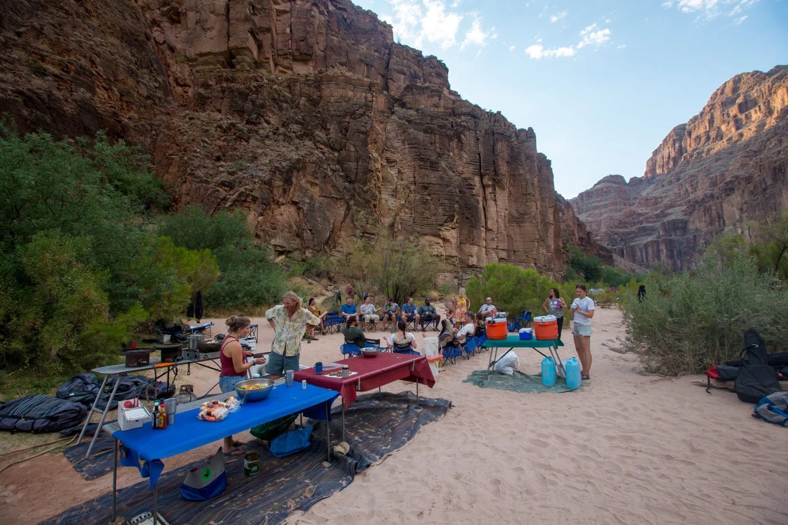 People gather around tables set up for a meal on a sandy riverbank, surrounded by canyon walls and greenery.