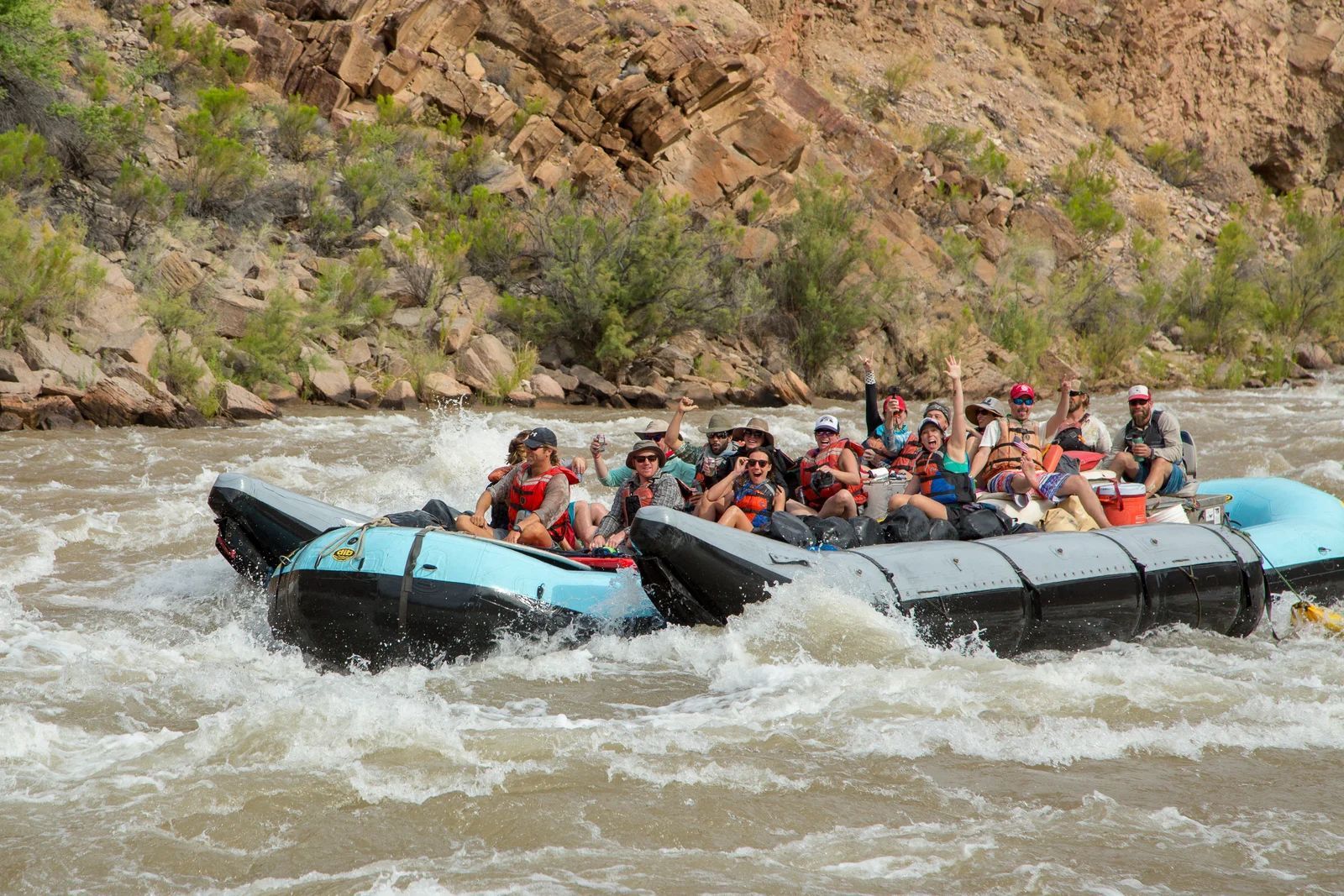 Group of people on rafts navigating a rapid, raising their arms, in a canyon.