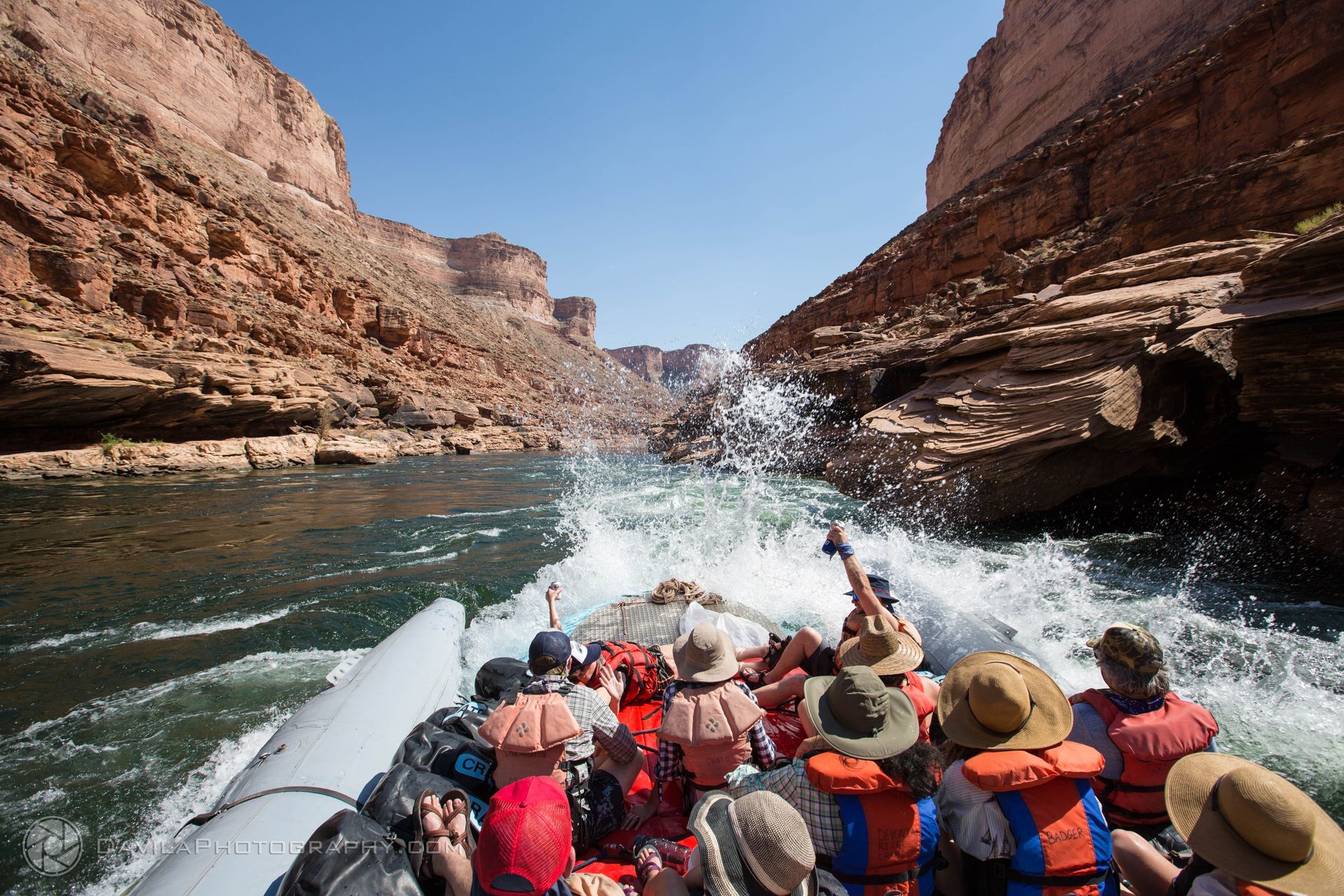 Rafting through a canyon, passengers in life vests get splashed. Rocky walls flank the water under a clear blue sky.