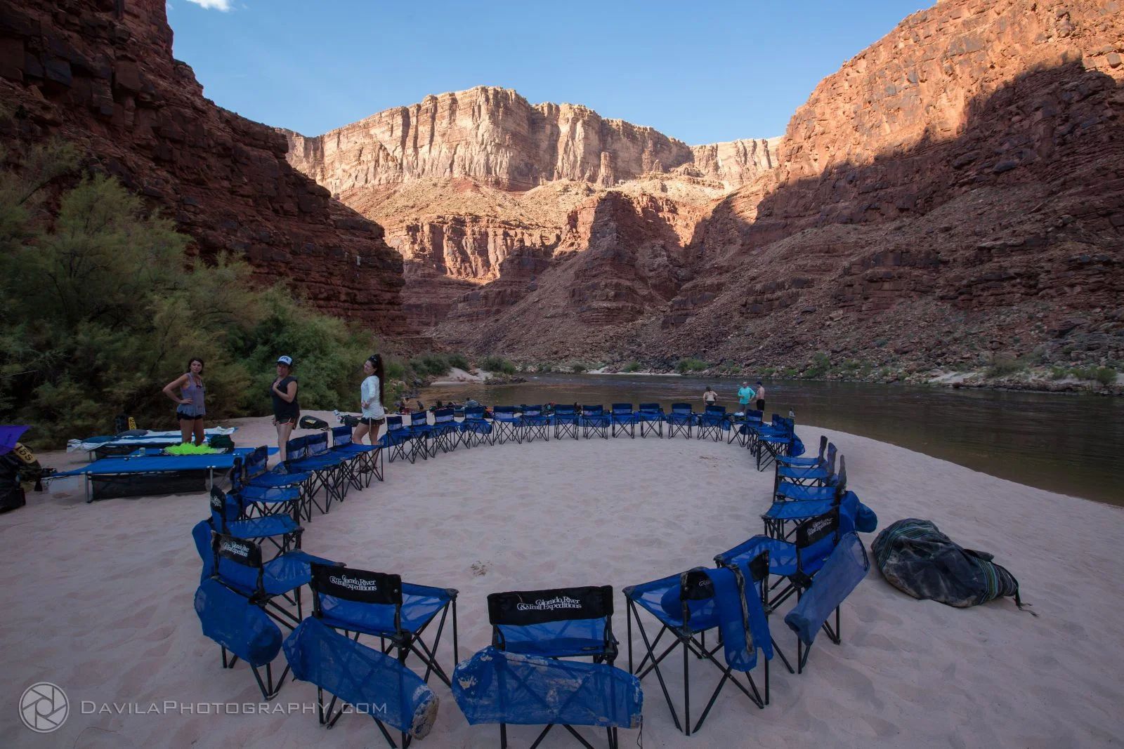 Circle of blue camp chairs on a sandy beach, with a river and canyon walls.
