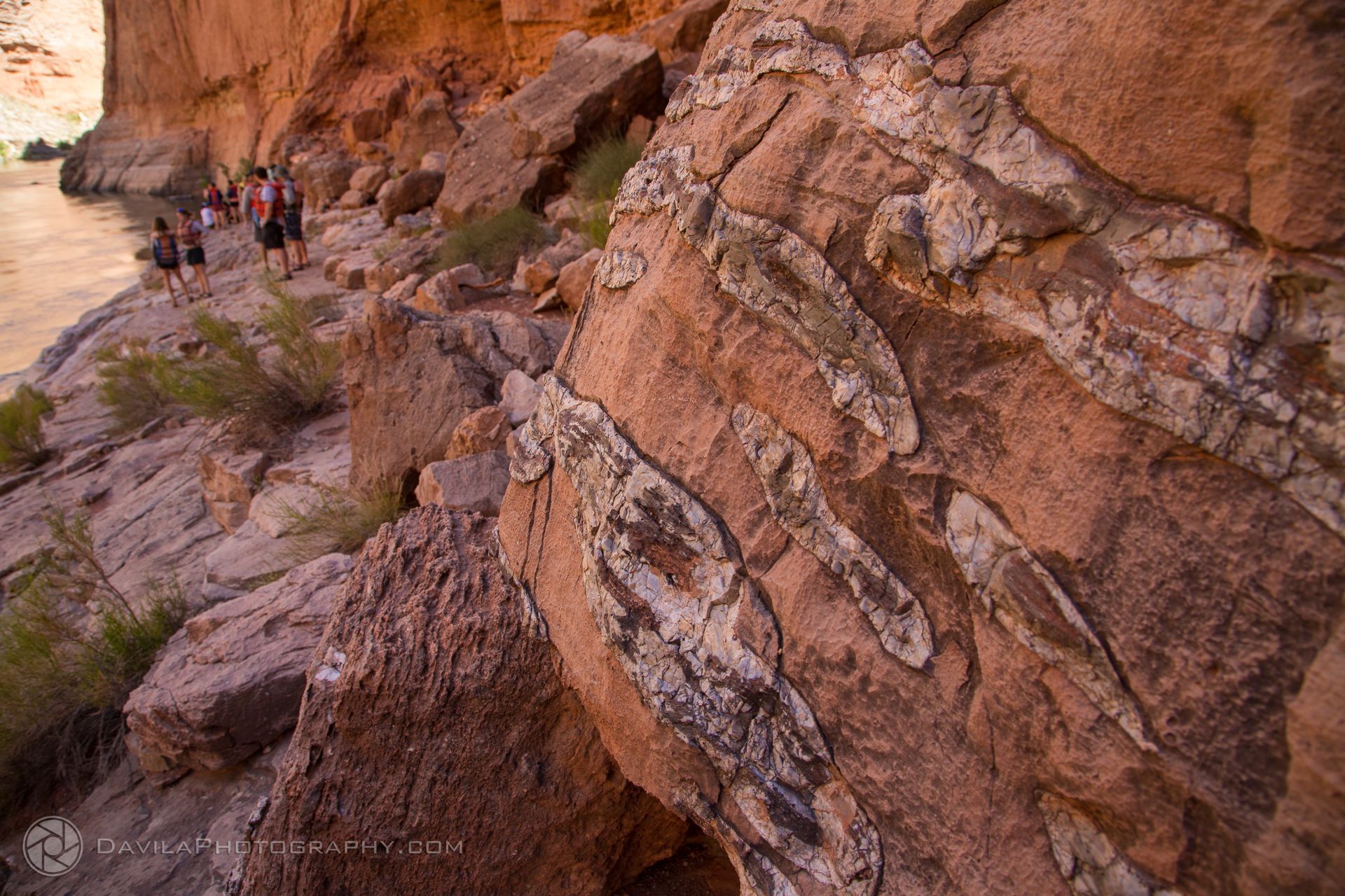 Red rocks with white veins, overlooking water, with a group of people standing nearby.