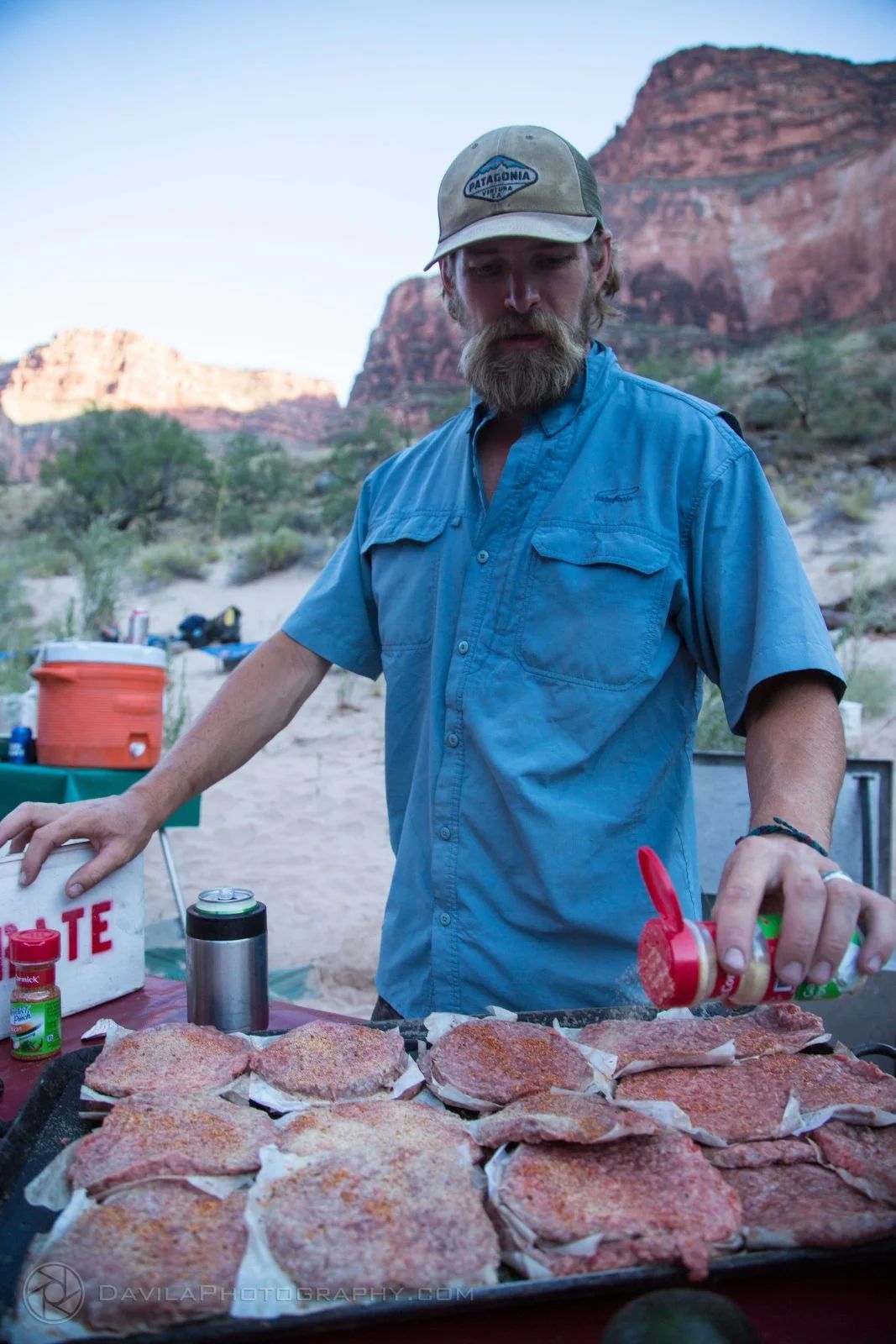Man seasoning meat patties on a grill outdoors, with a canyon backdrop.