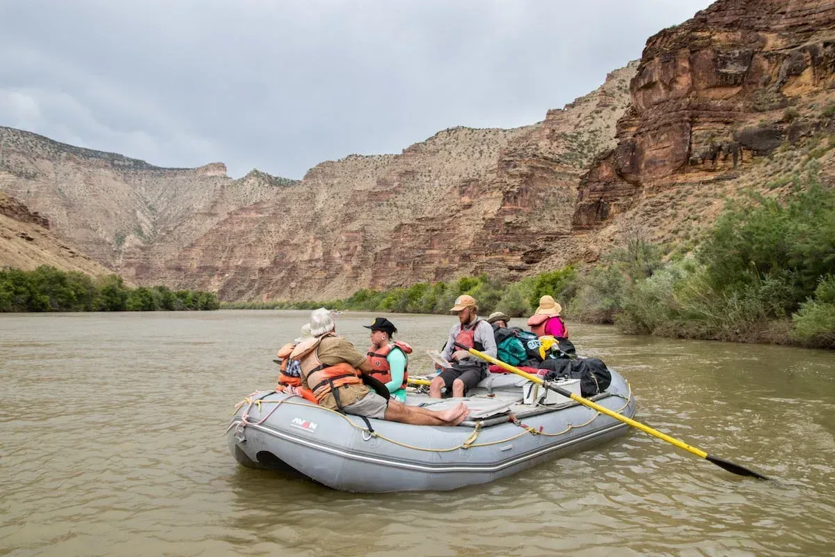 Rafting on a brown river through a canyon. Four people wearing life vests, mountains in background.