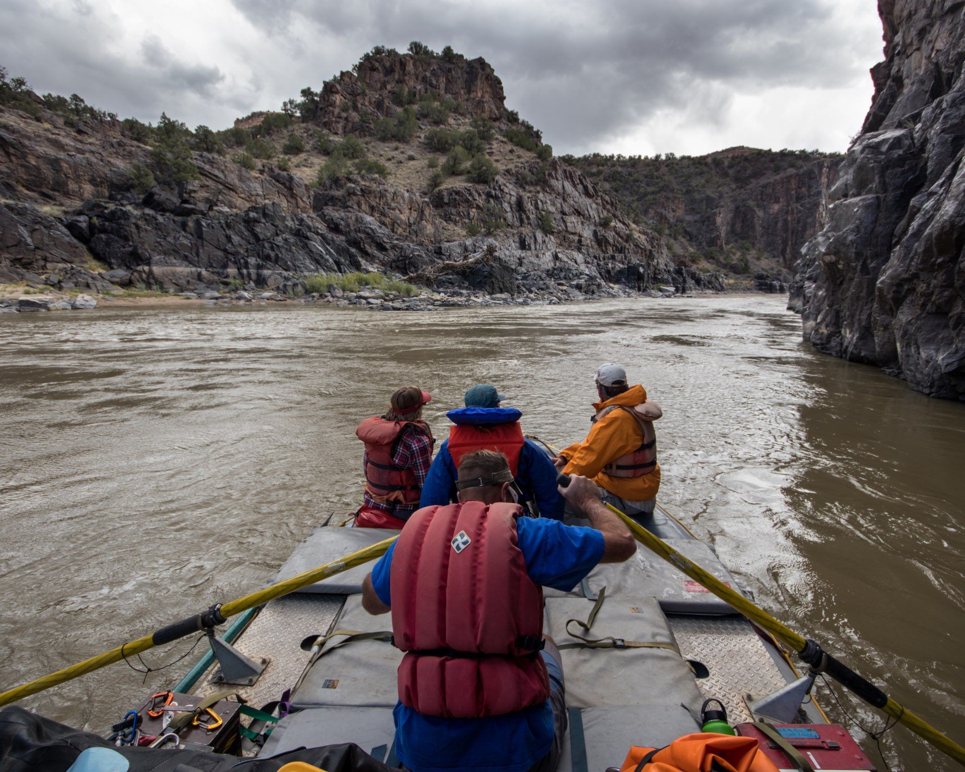 People in a raft on a river, navigating through a canyon with rocky cliffs under a cloudy sky.