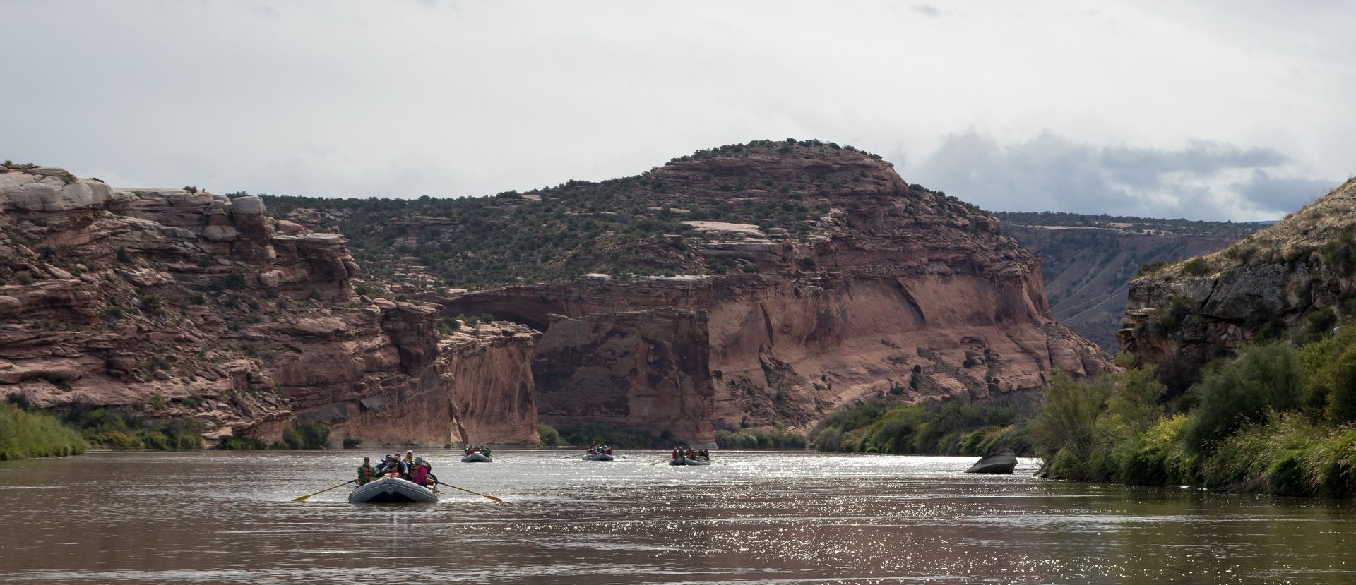 Rafts on a wide river with rocky cliffs on either side. Cloudy sky.