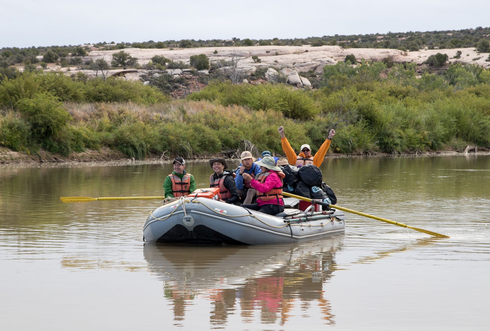 People on a raft with paddles on a river, arms raised in excitement. Green vegetation and tan hills in the background.