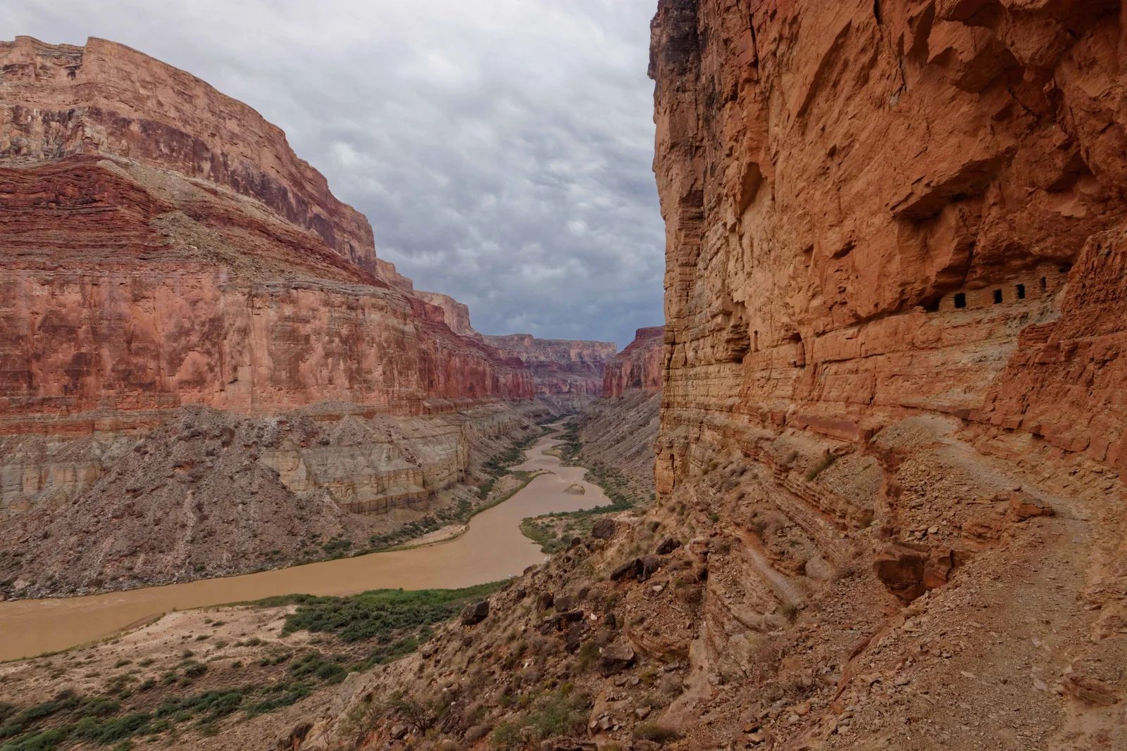 Canyon landscape with reddish-brown rock walls, a muddy river, and cloudy sky.
