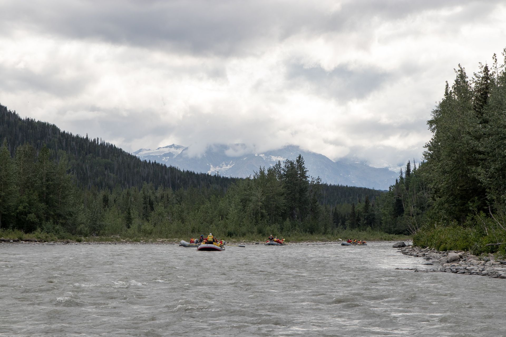 Rafts on a river in a forested valley with mountains in the background under a cloudy sky.