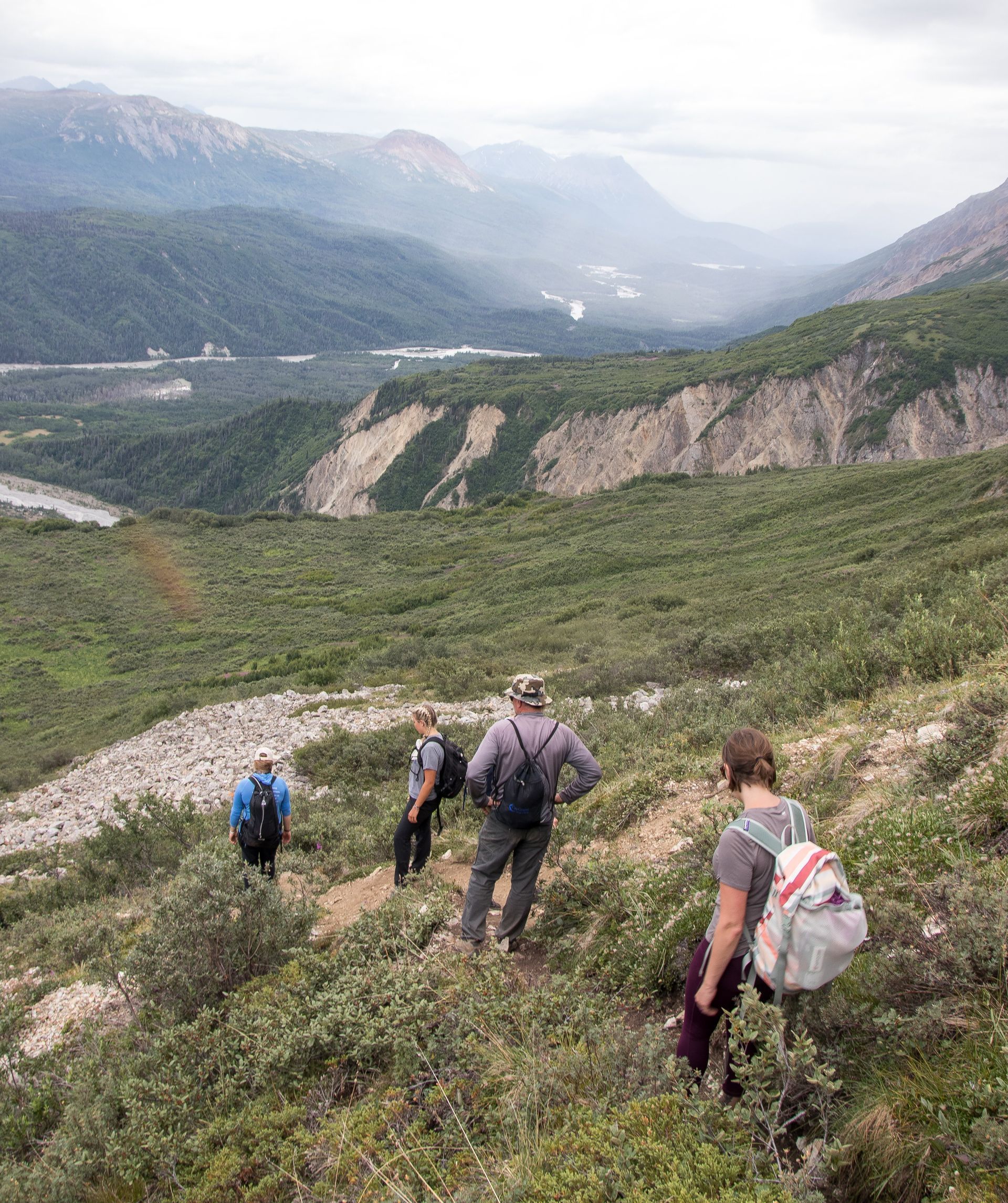 Hikers descend a grassy hillside toward a river valley, mountains in the distance.