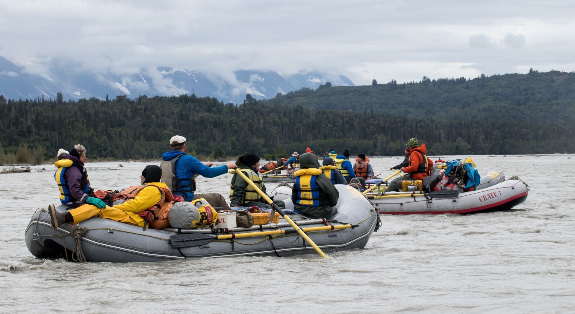 Rafts of people on a river. Cloudy sky, mountains, and trees in the background.