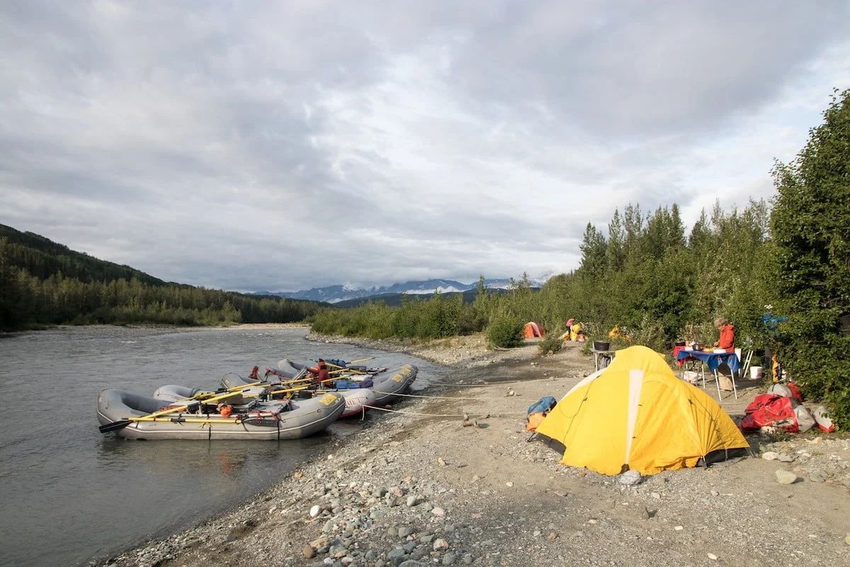 Yellow tent and rafts on a riverbank, people are camping. Mountains in the distance under a cloudy sky.