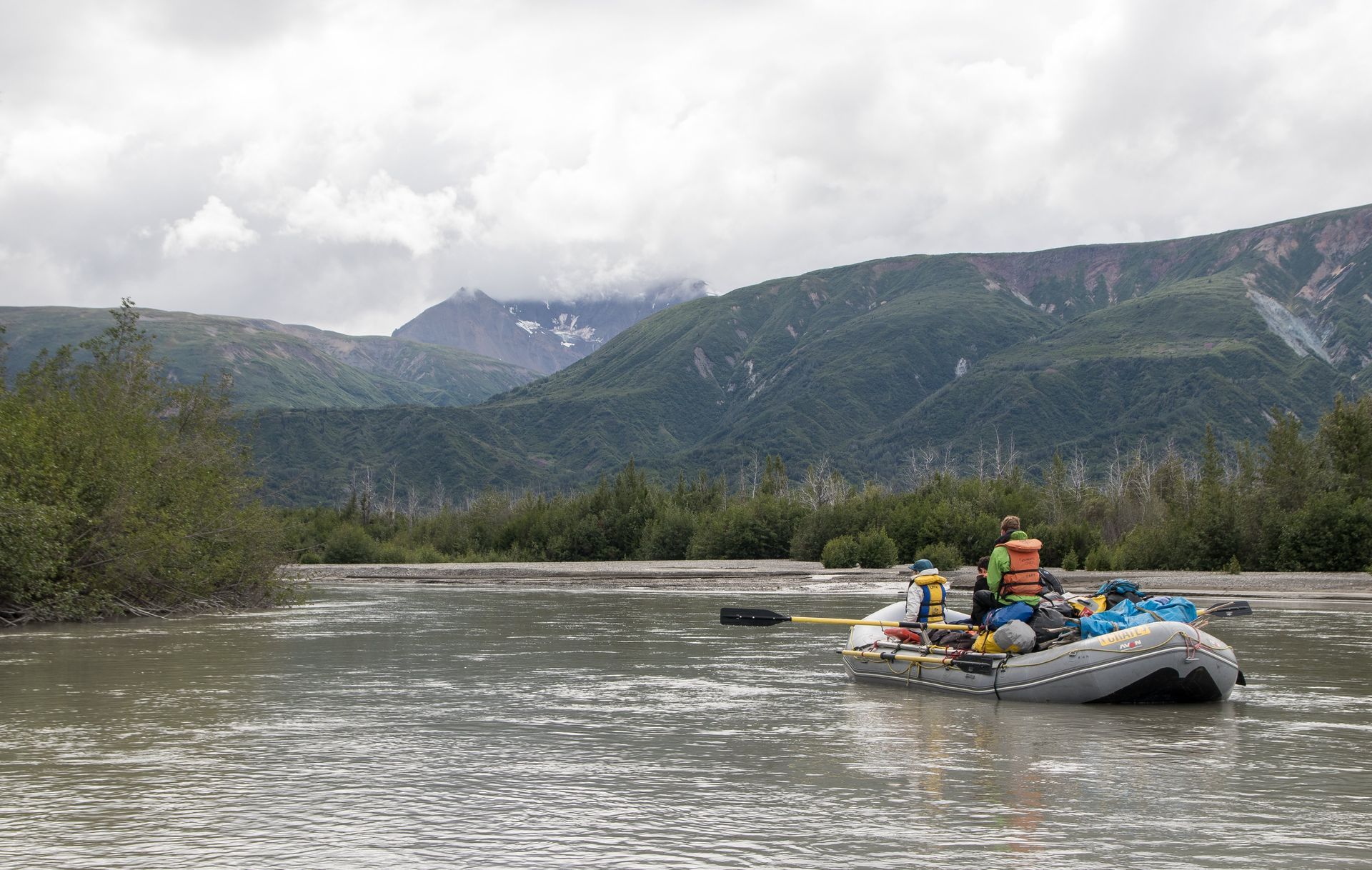 Raft with people on a river, mountains in the background under a cloudy sky.