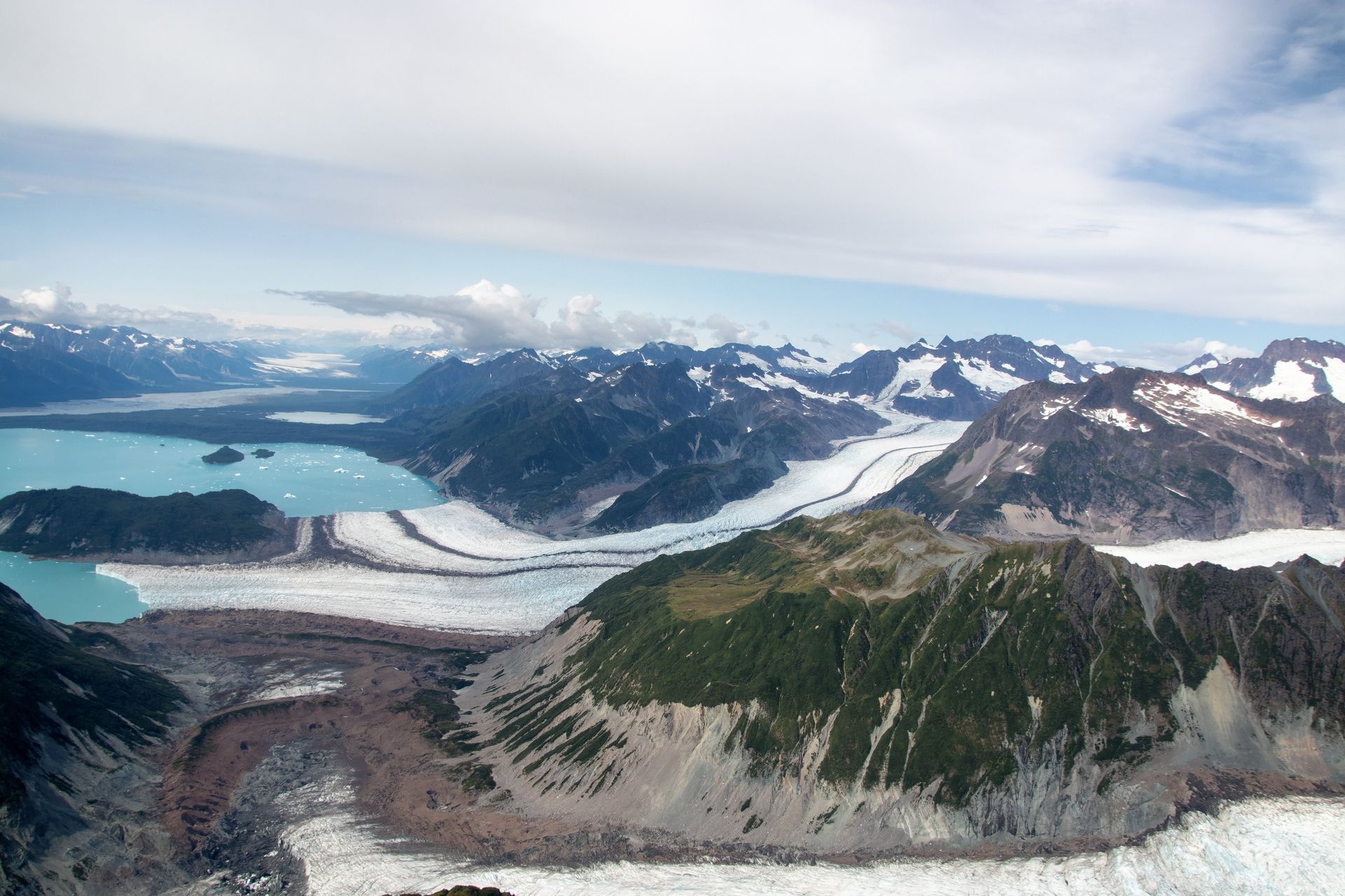 Aerial view of glacial landscape; glaciers, turquoise lakes, mountains under cloudy sky.