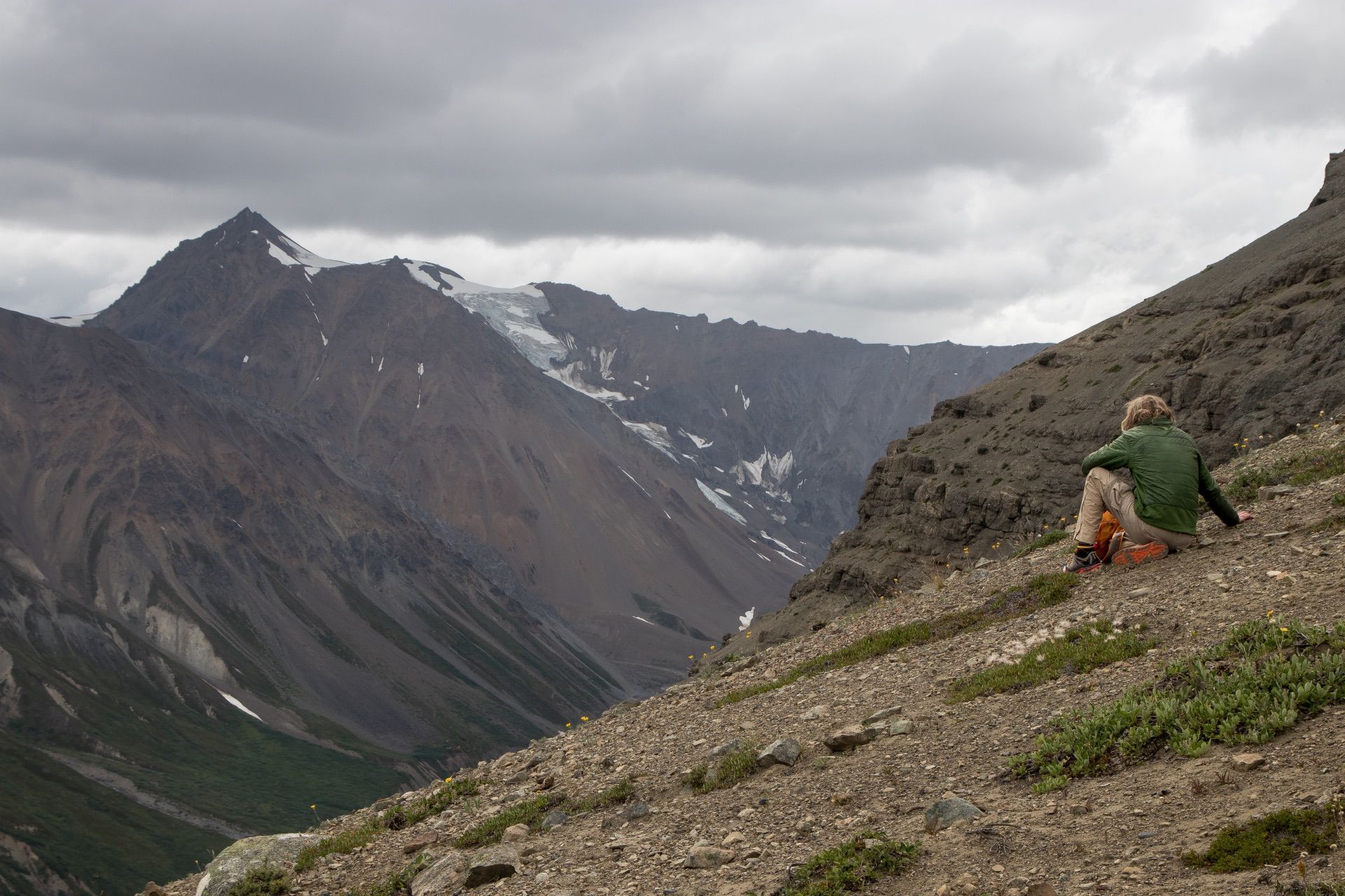 Person rests on a rocky mountain slope, overlooking a valley with mountains under a cloudy sky.