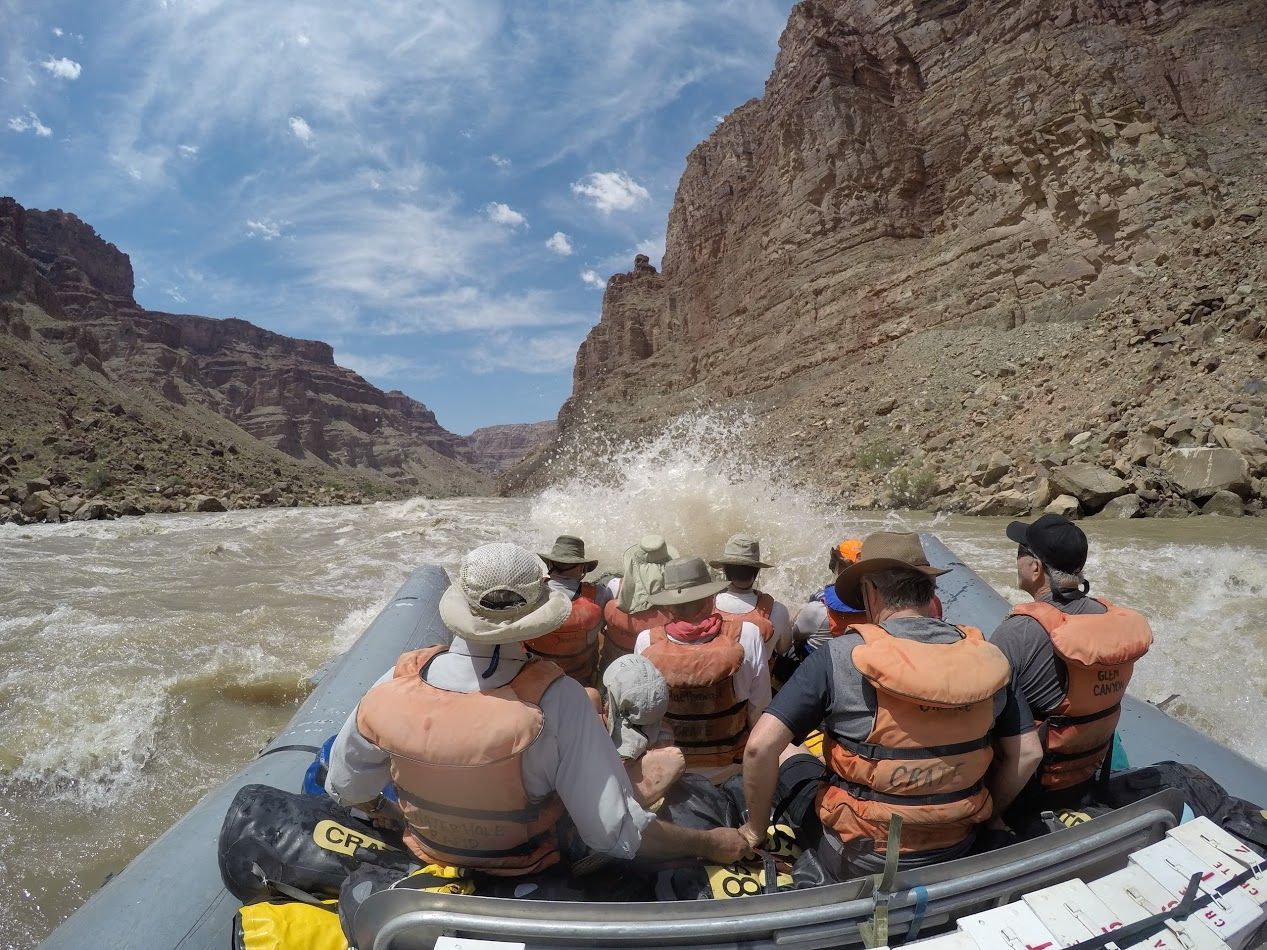 Rafting through a canyon, passengers in life vests get splashed by whitewater.