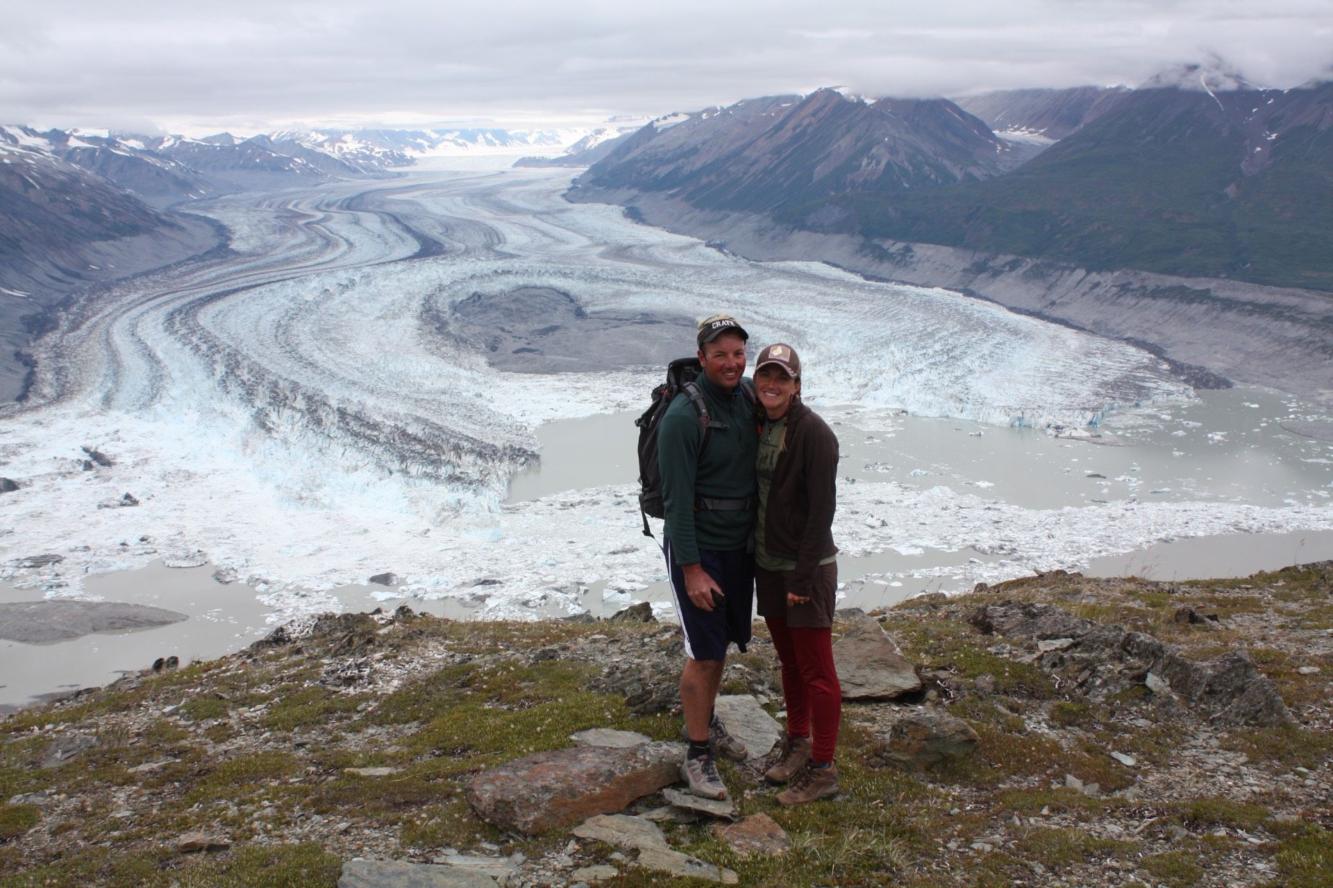 Couple standing on a mountain overlooking a large glacier. Mountains and overcast sky in background.