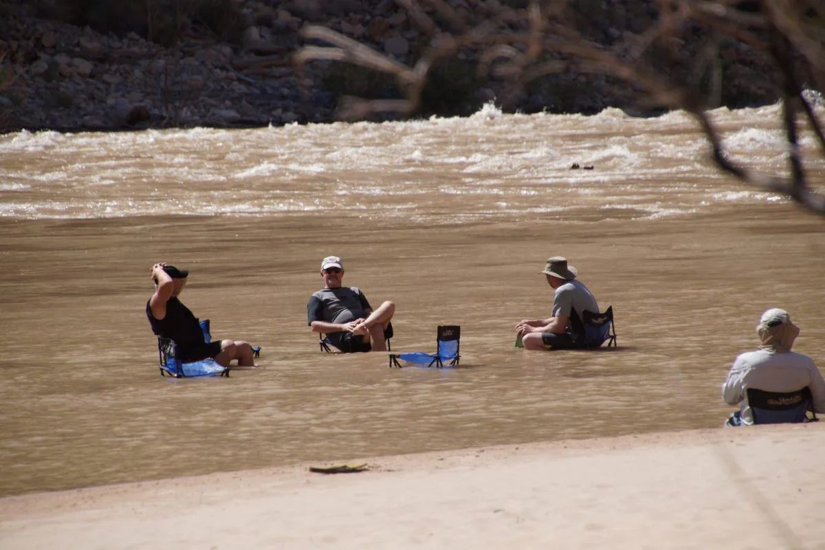 Four people sitting in folding chairs in shallow, muddy water near a flowing river.