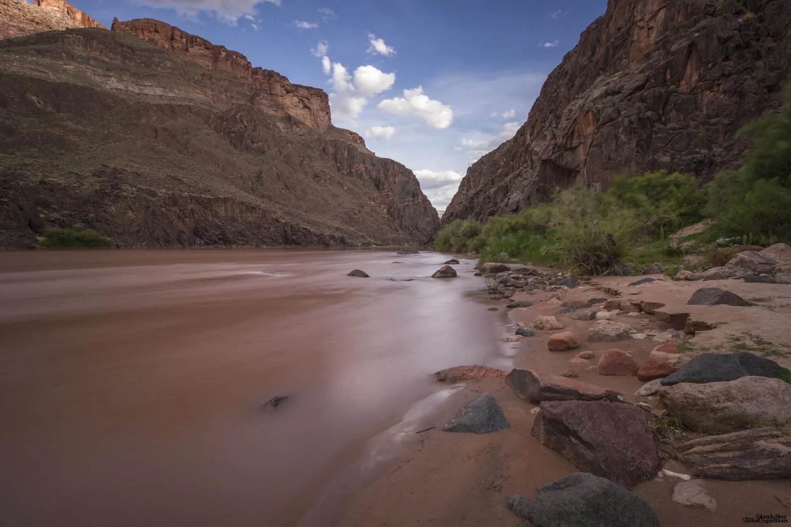 Brown river flows through a canyon, sandy bank with rocks on the right. Rocky canyon walls and blue sky.