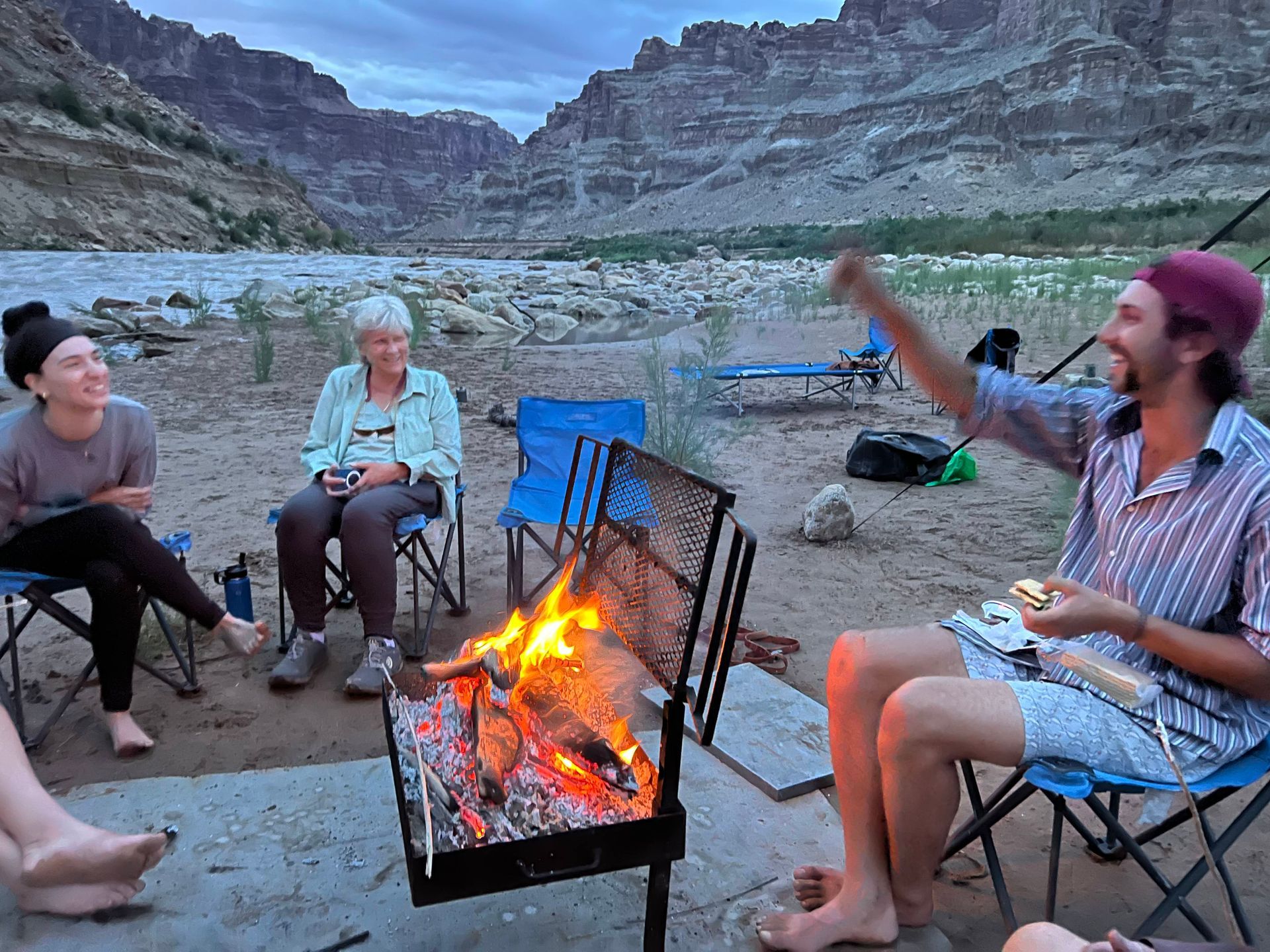 People by campfire on riverbank, grilling food. Rocky canyon in background.