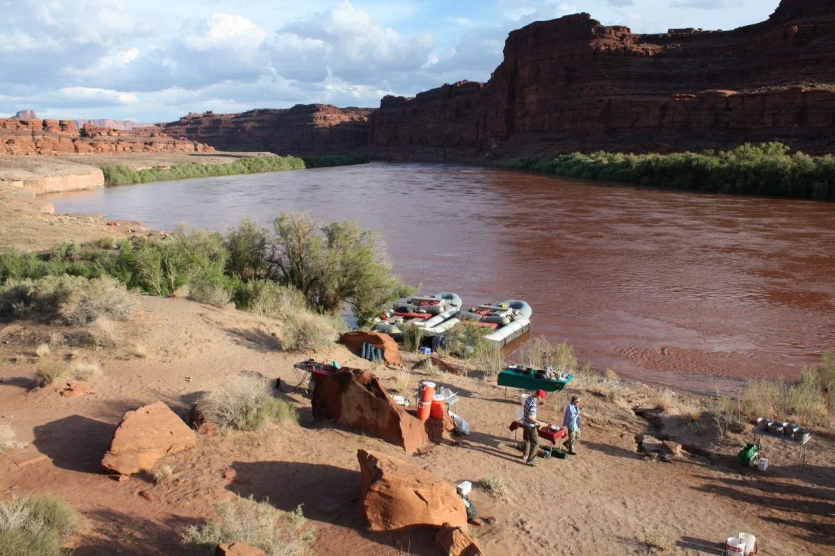 Campsite on sandy riverbank next to red river. Two rafts, red rock cliffs in background, sunny.