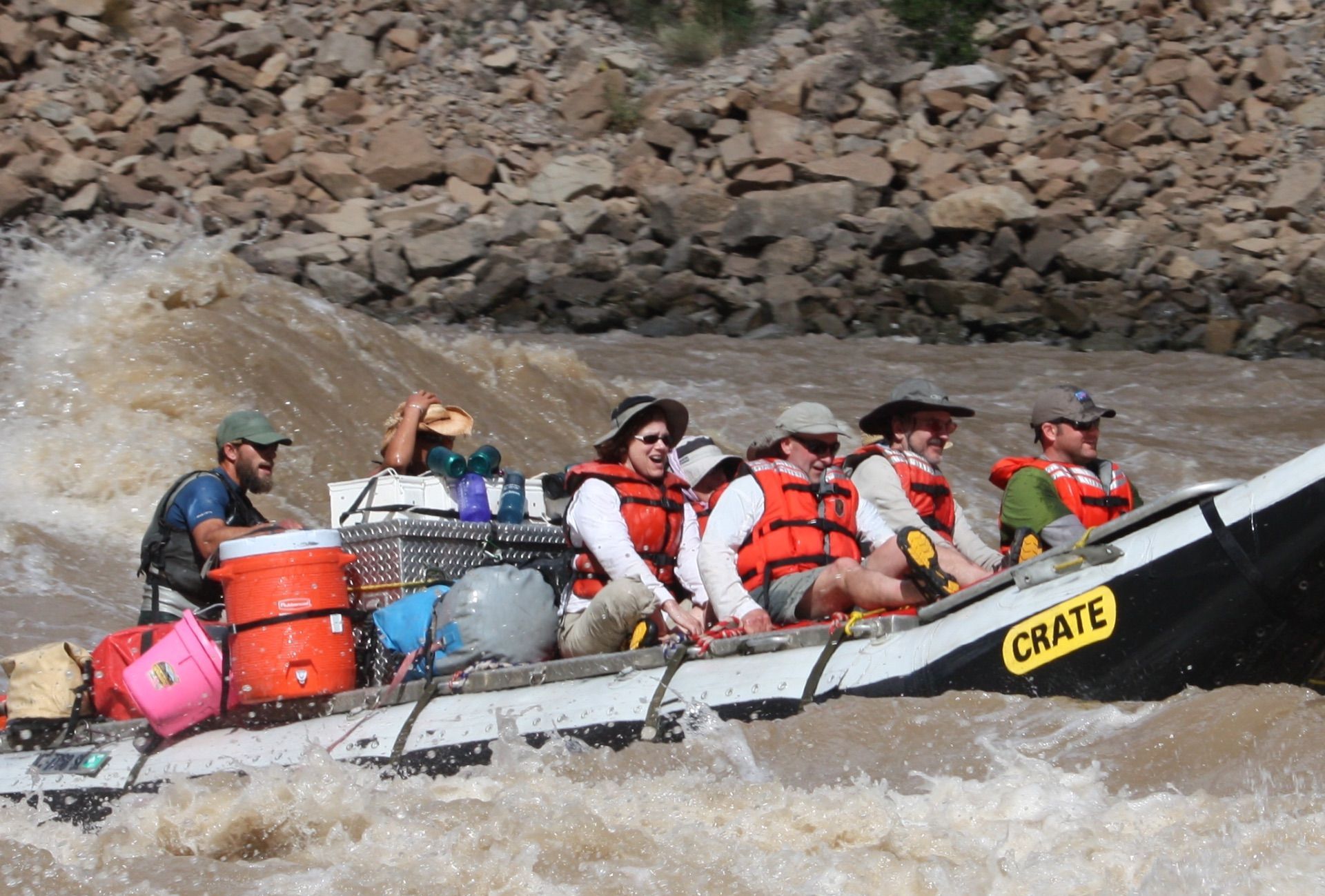 A raft with six people navigates a river. The boat is packed with gear and moves through rapids.