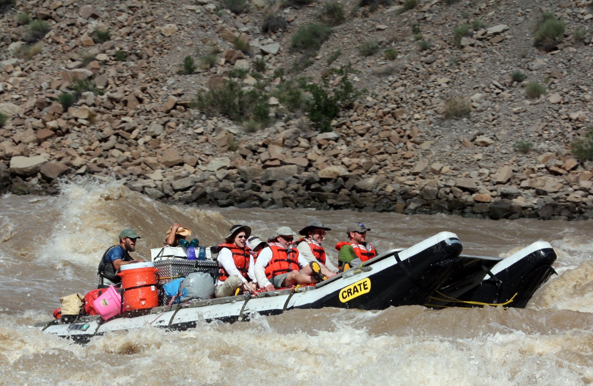 Raft with people navigating a whitewater rapid; rocky canyon walls in background.