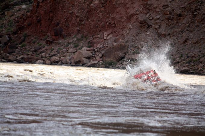 A raft capsizes in a turbulent river, spraying water against a red-brown rock face.