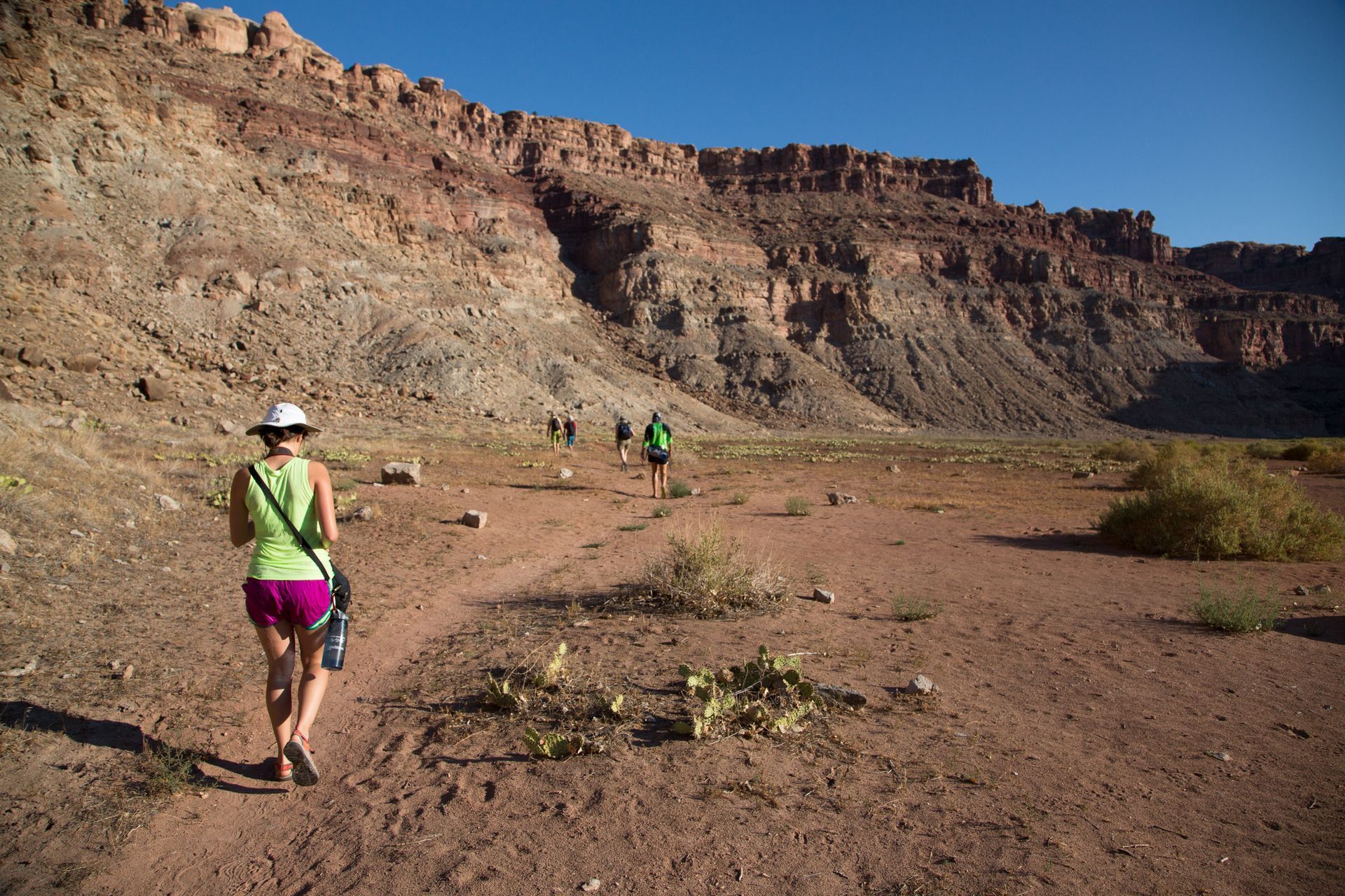Hikers walk on a dirt path towards a red rock canyon under a blue sky.