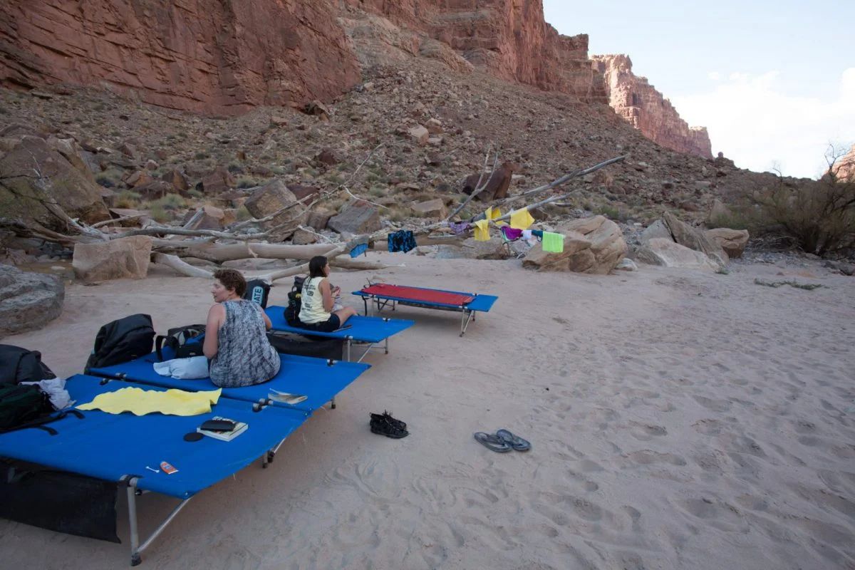 Two people relax on blue cots at a sandy campsite, near a river and red rock cliffs.