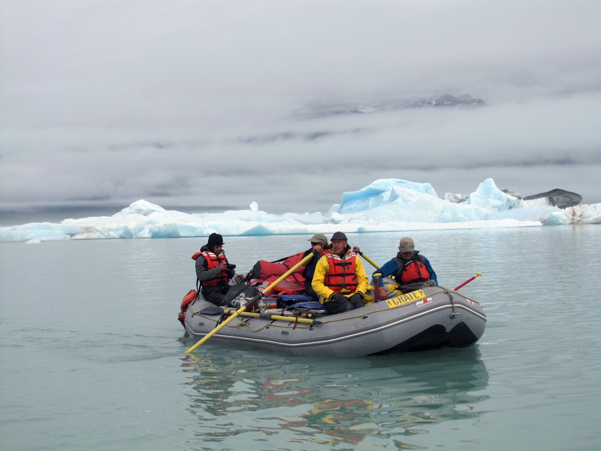 People in a raft with oars near icebergs, cloudy day.