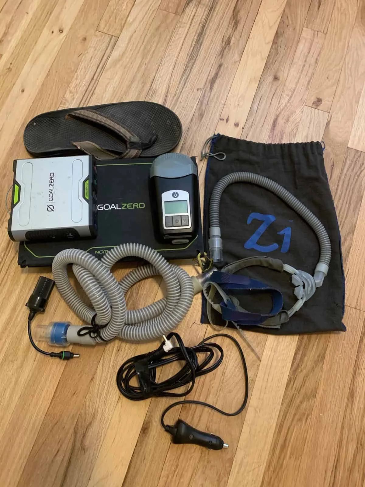 Items on wooden floor: CPAP machine, battery, mask, tubing, sandals.