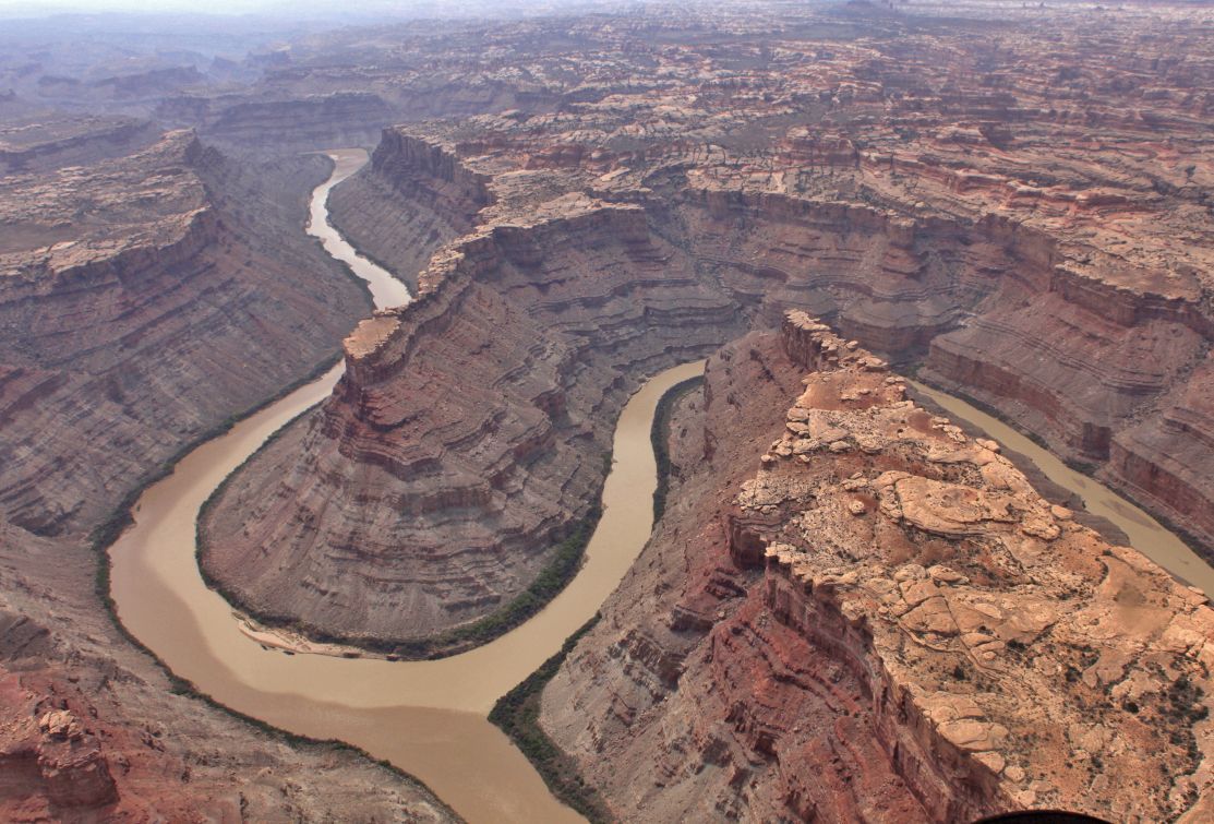 Aerial view of a brown river winding through a sandstone canyon in an arid landscape.