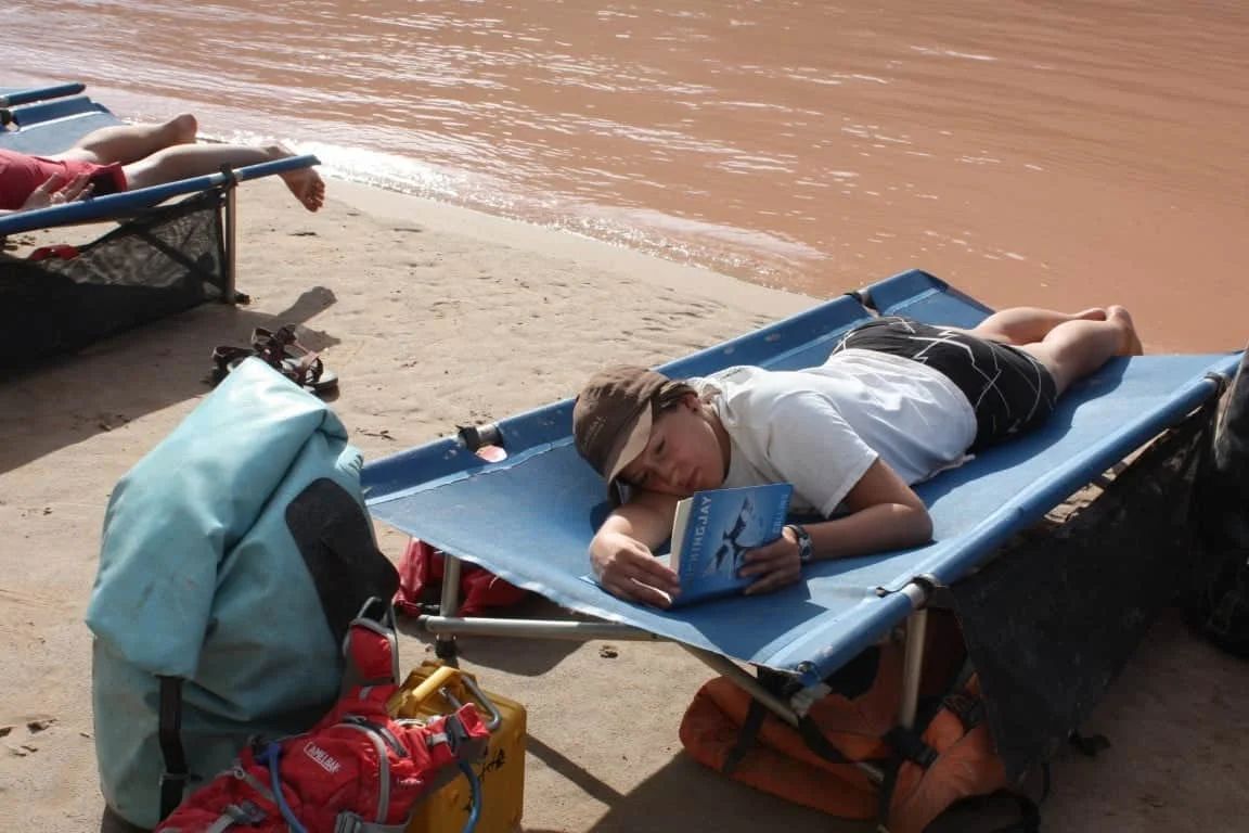 Person lying on a blue cot, reading a book on a sandy beach next to a river.