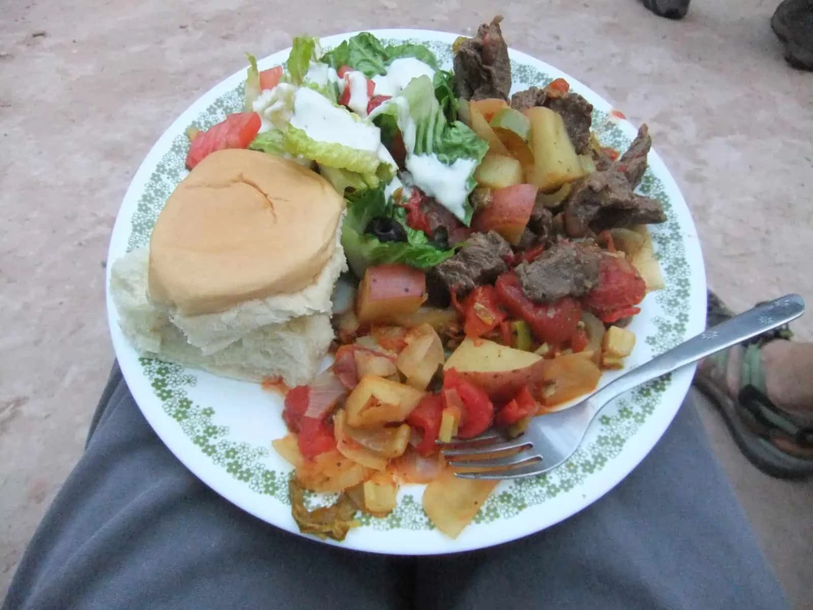 Plate with meat, vegetables, bread, and salad; person's legs and shoe in frame.