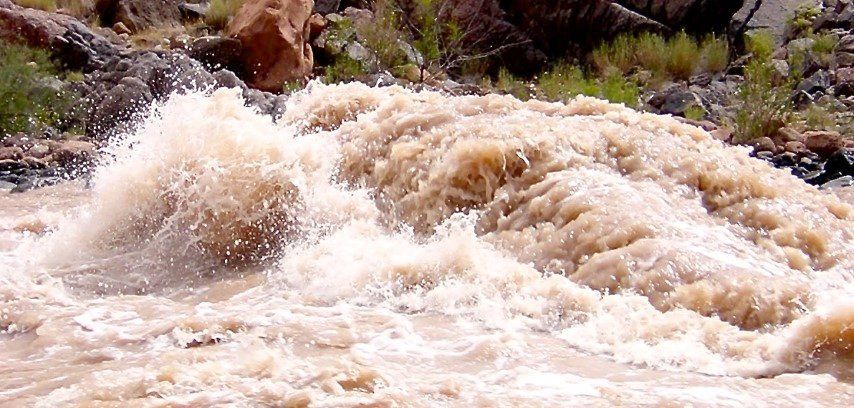 Fast-flowing muddy river with white splashes and brown rocks in the background.