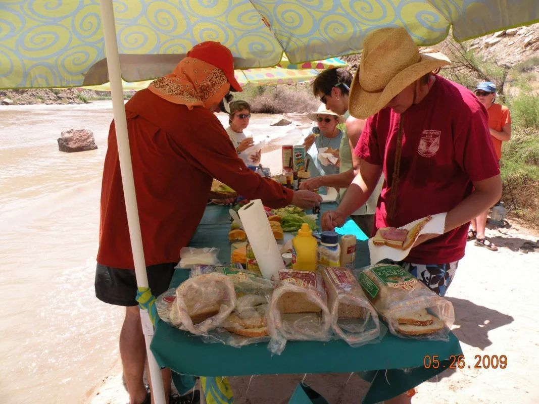 People making sandwiches at a riverside picnic table, shaded by an umbrella.