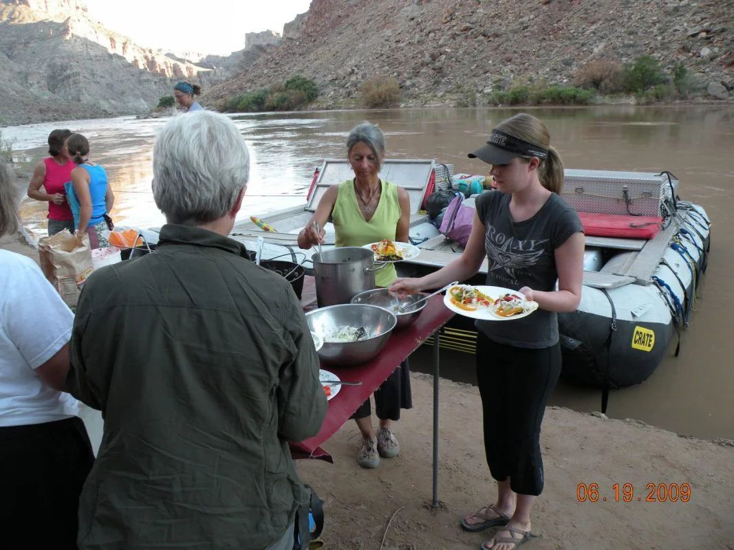 People serving food from a riverside table near a raft.