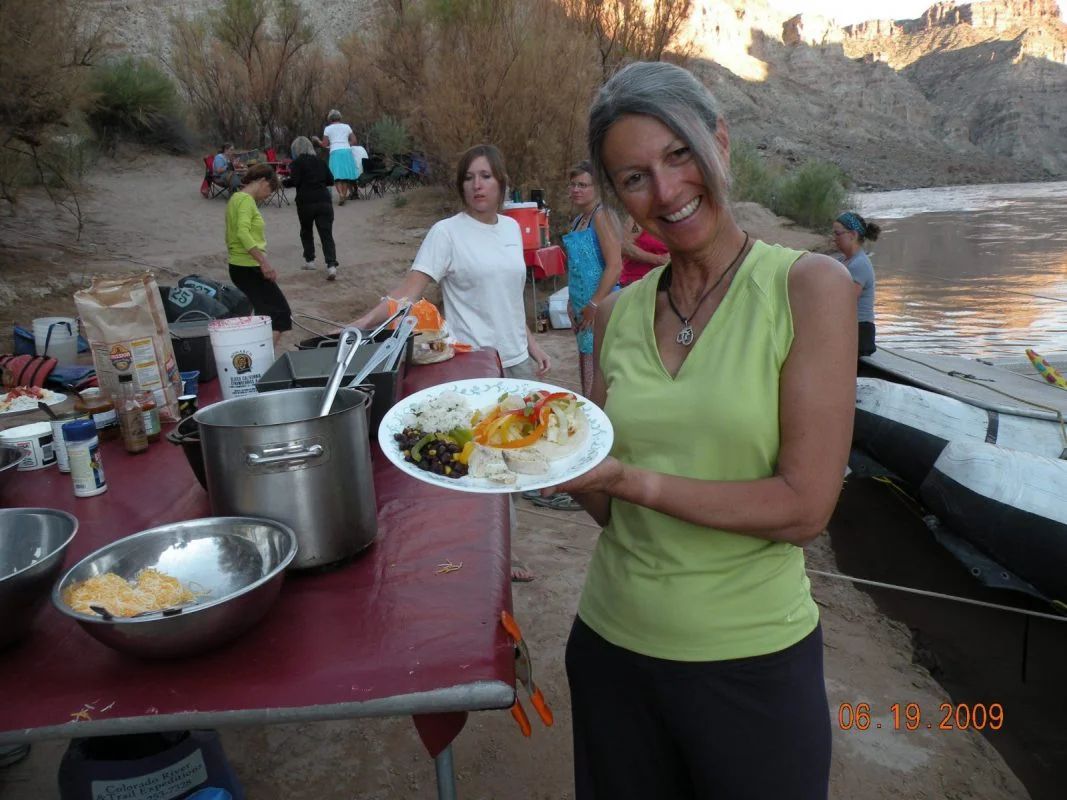 Woman smiling, holding plate of food at riverside picnic. Others nearby, cooking and eating.