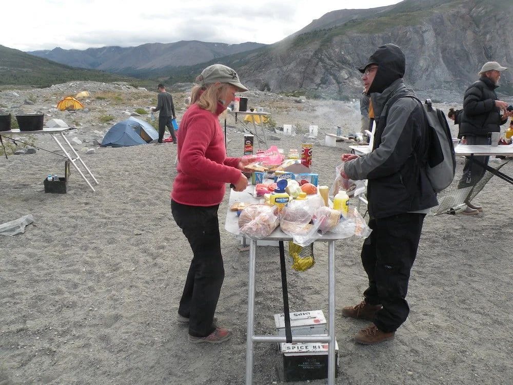 Woman and man at a table with food, likely a camp, in a mountainous, rocky setting.