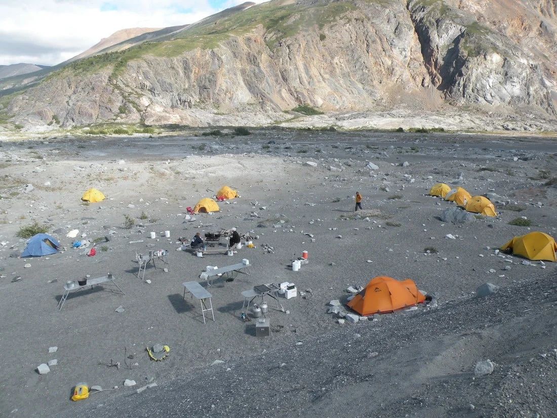 Campsite with several tents set up on a rocky, gray terrain near a mountain, person standing.