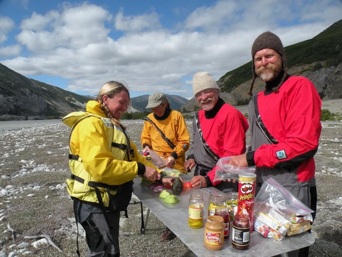 Group of people preparing a meal outdoors next to a river. They are wearing rain gear.