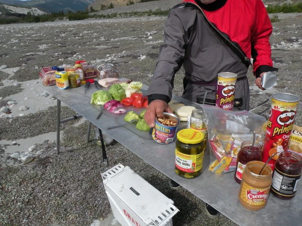 Person in red jacket prepares a meal outdoors on a metal table, with various food items including Pringles and canned goods.