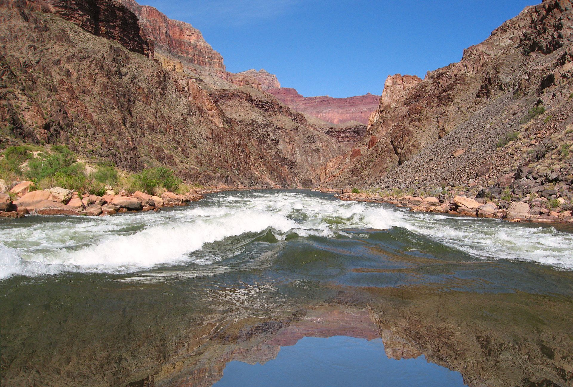 White water rapids in a canyon, with red rock walls and blue sky reflected in the water.