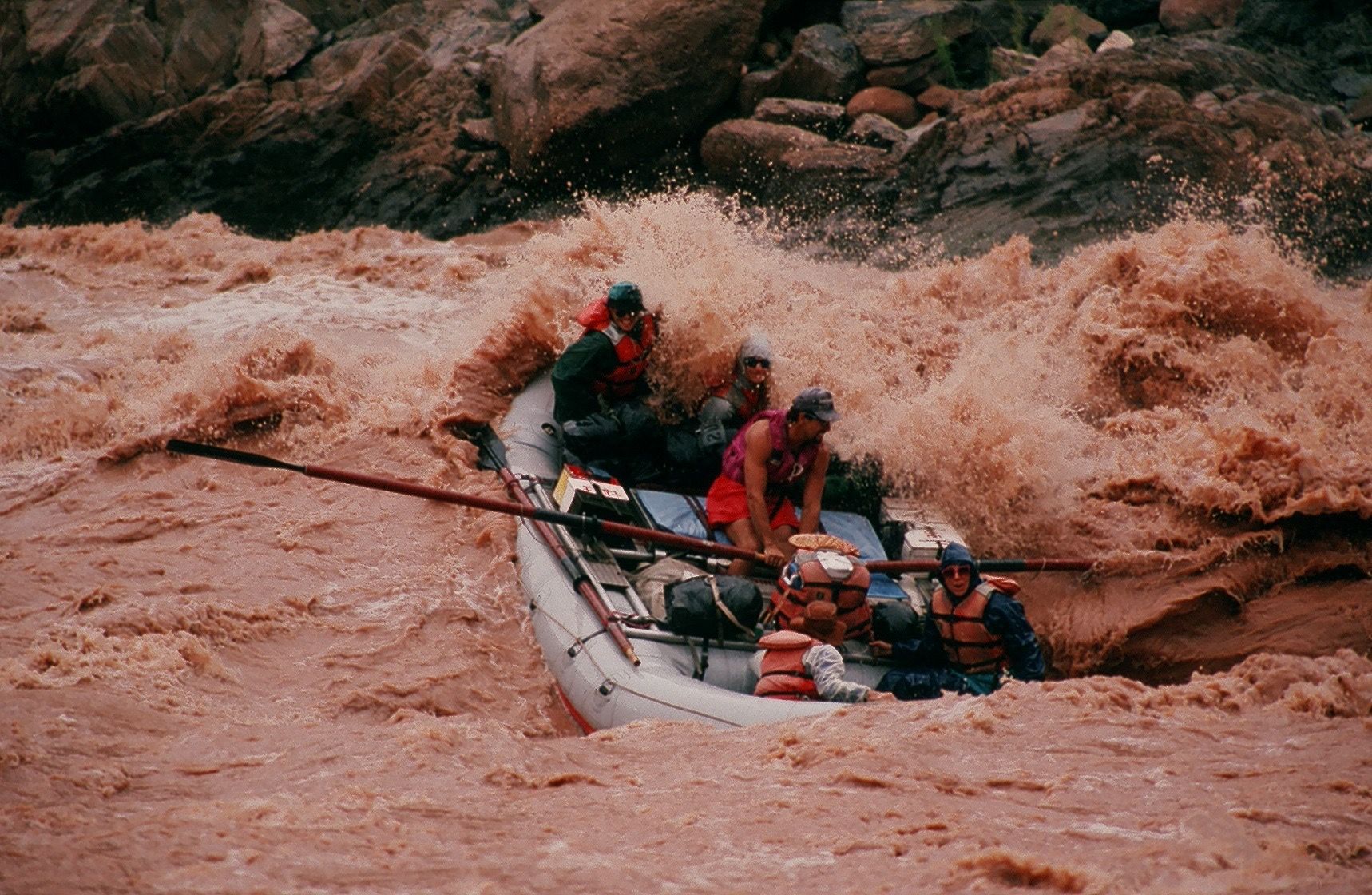 Rafting through churning, muddy water. Several people in a boat navigating a rapid.