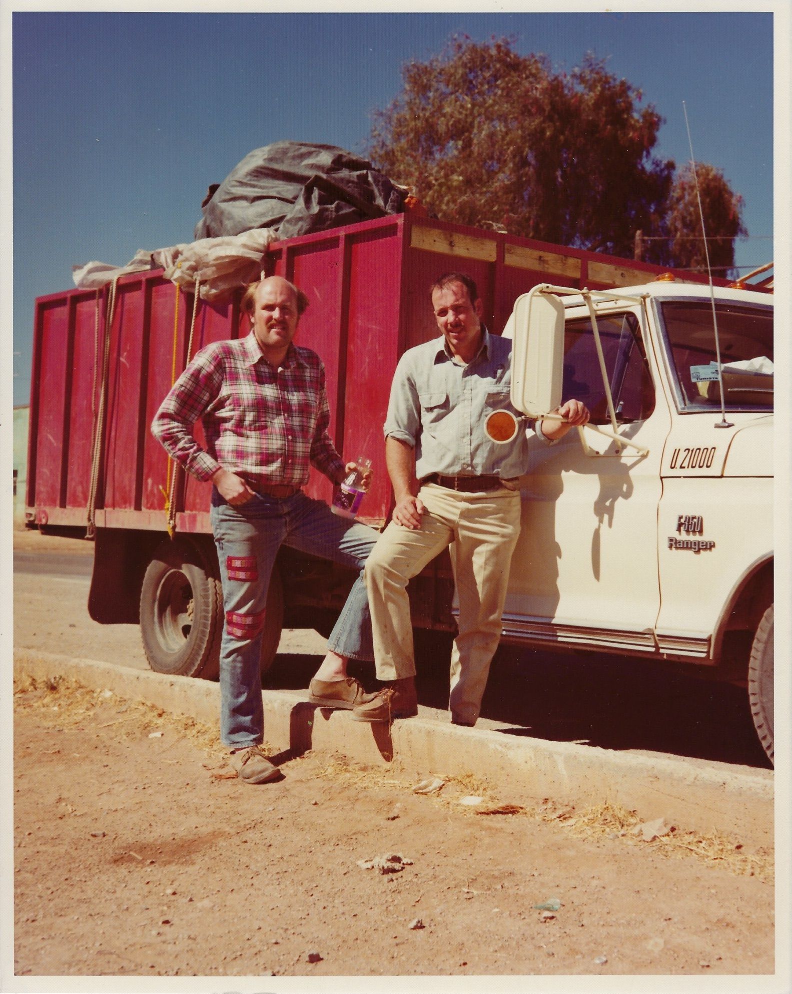 Two men pose by a white truck with a red cargo bed. One man wears jeans, the other khaki pants. Desert setting.