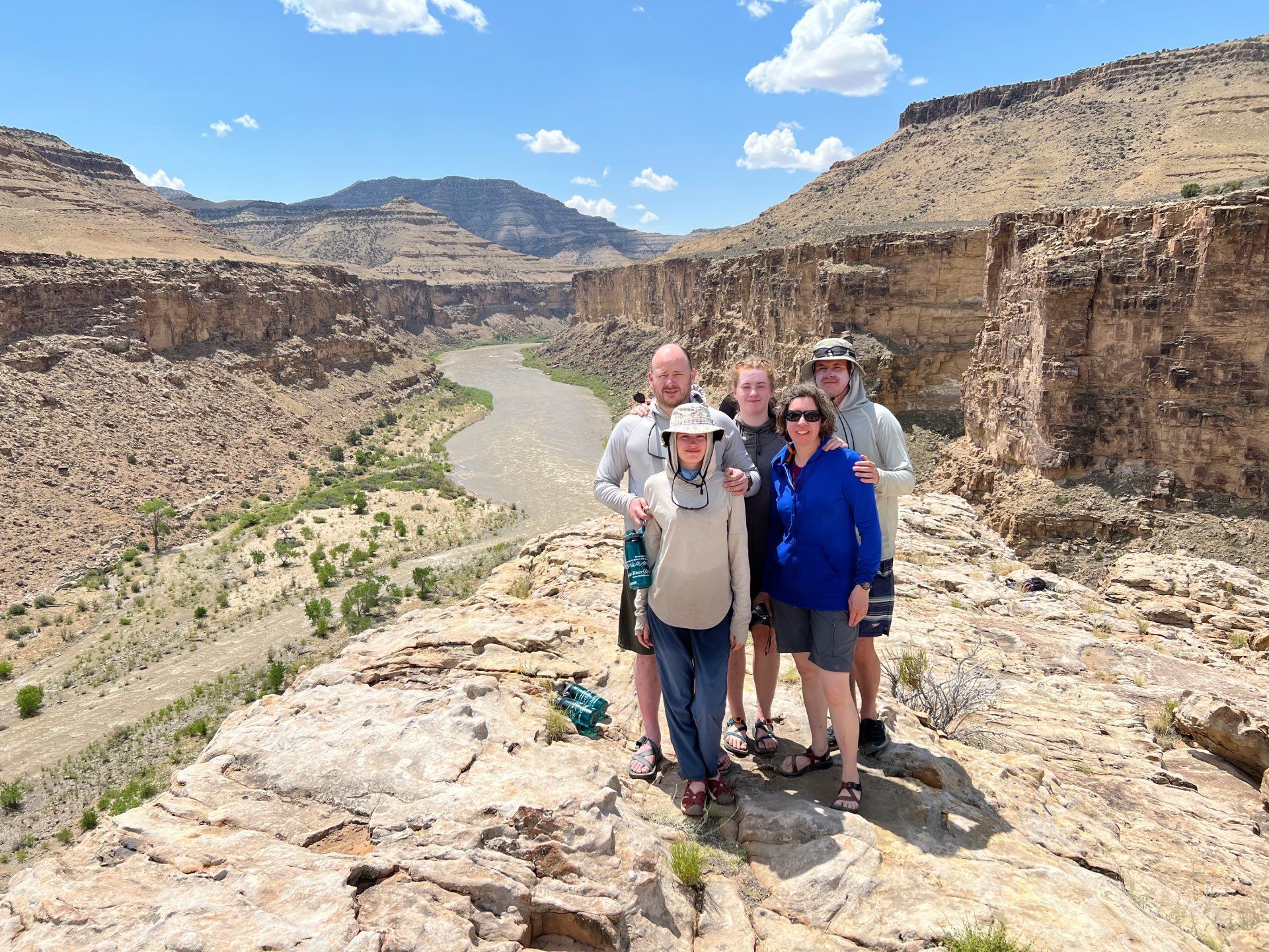 Group poses on a cliff overlooking a canyon with a river. Sunny, blue sky.