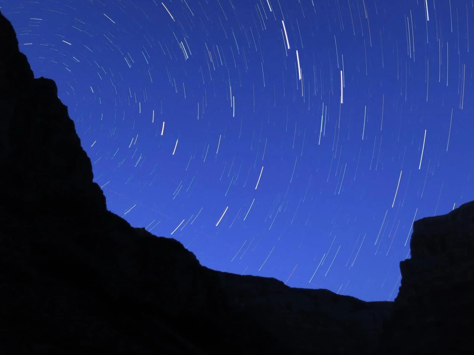 Night sky with star trails seen through a silhouetted canyon. Blue and white colors dominate the scene.