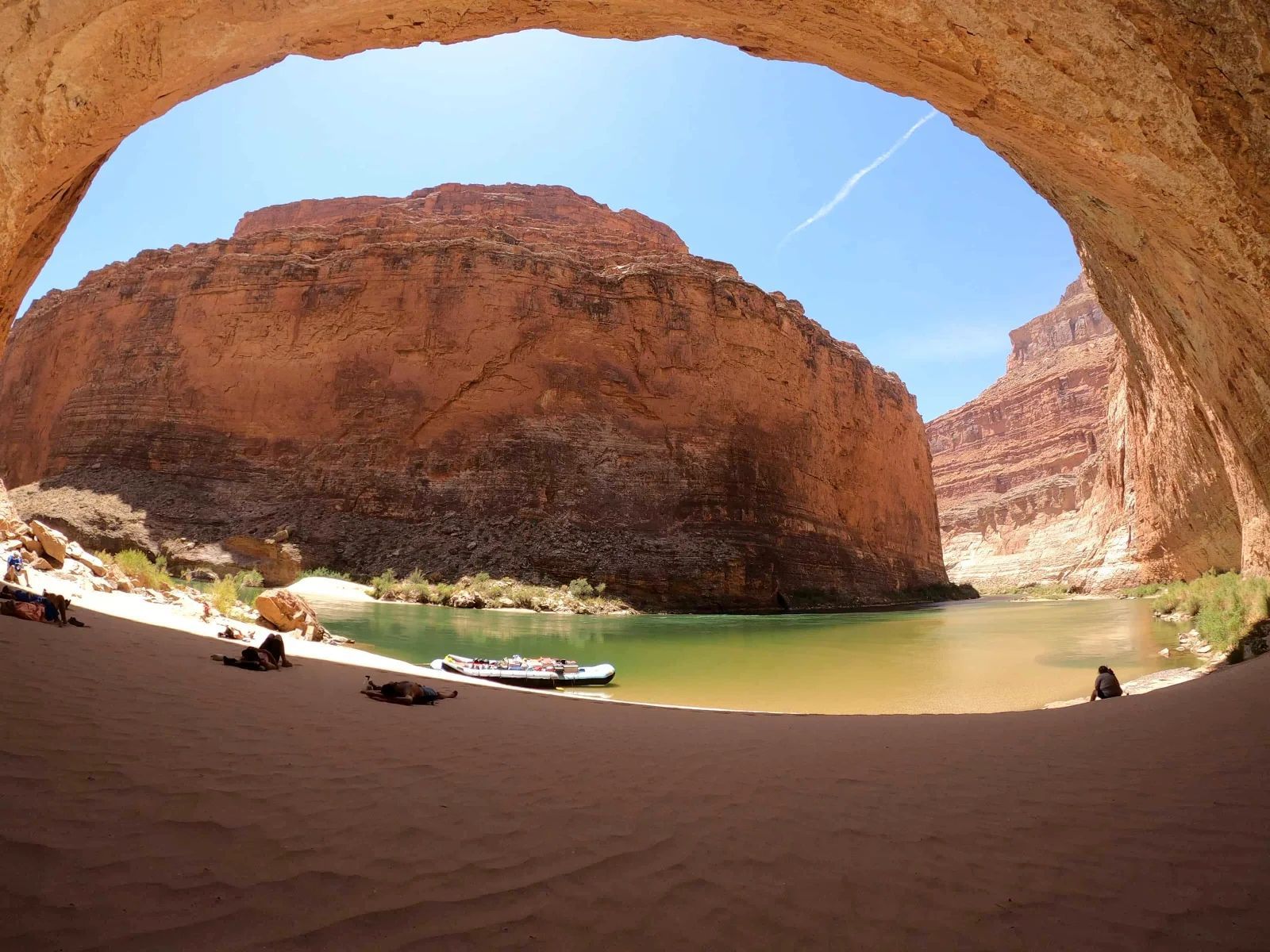 Sandy beach inside a cavern overlooks a river, with red rock canyon walls and a blue sky.